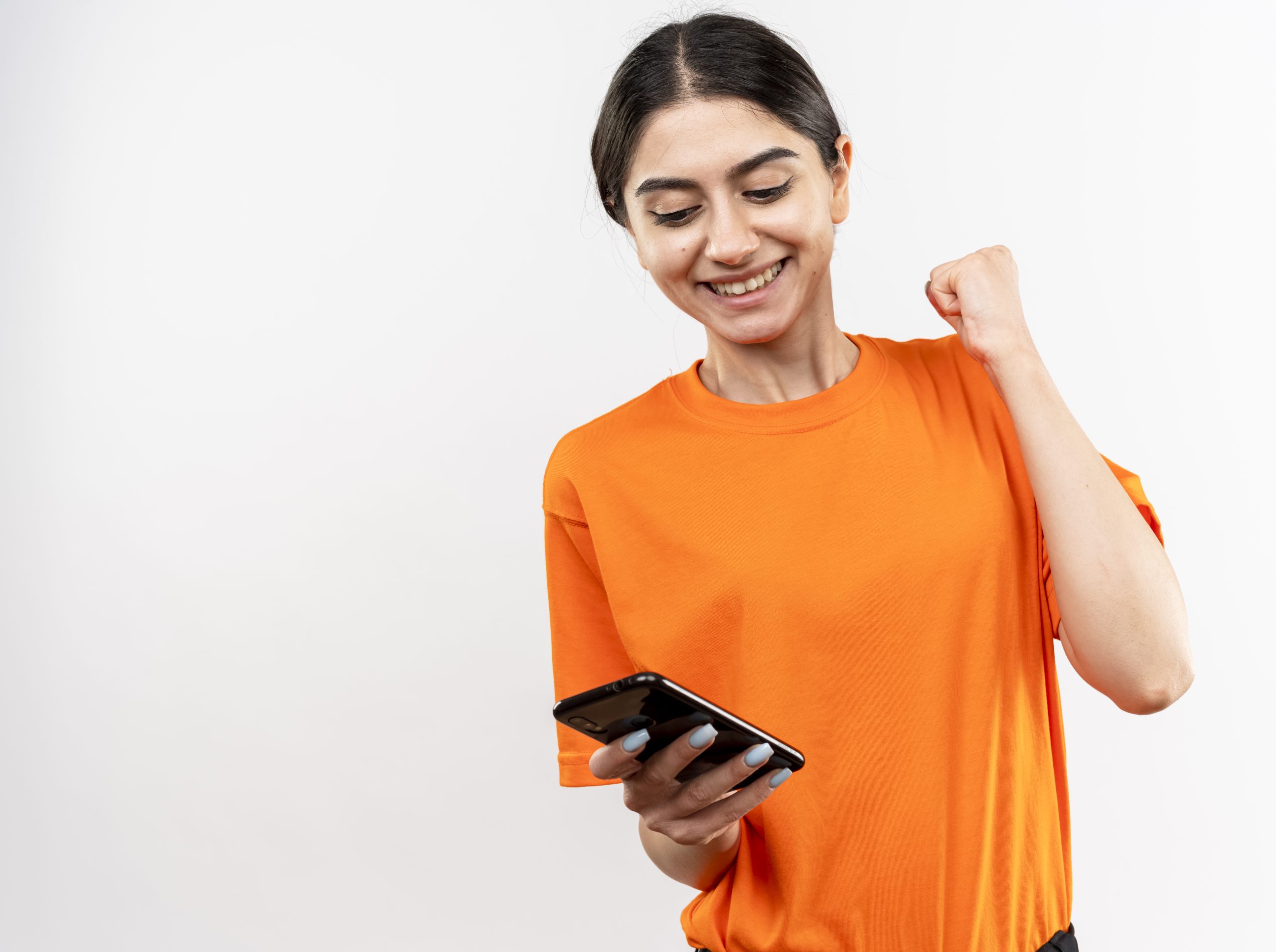 Young Girl Wearing Orange T Shirt Holding Smartphone Clenching Fist Happy Excited Smiling Cheerfully Rejoicing Her Successstanding White Wall Scaled
