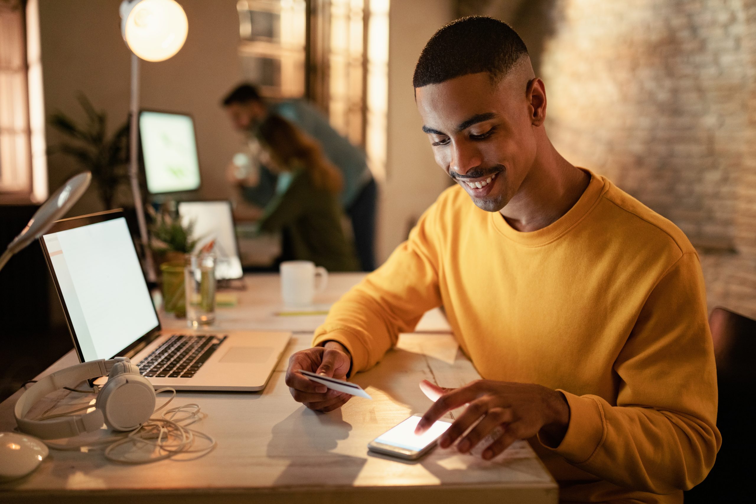 Happy African American Entrepreneur Using Mobile Phone Credit Card From Online Payments While Working Night Office Scaled